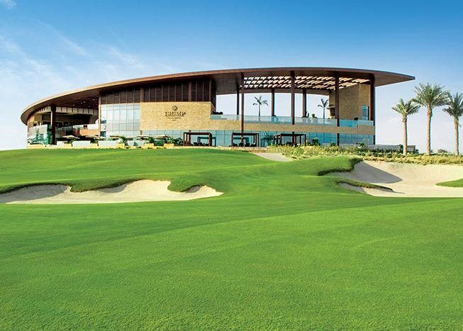 Club-house de golf moderne avec de grandes fenêtres en verre et un toit incurvé à Damac Hills, surplombant un parcours de golf bien entretenu avec des bunkers de sable et de l'herbe verte.