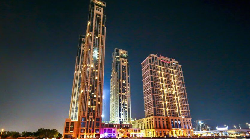 An animated nighttime cityscape showing three tall buildings, including the Amna Tower, illuminated by colored lights with reflections on a calm body of water in the foreground in the city of Al Habtoor.