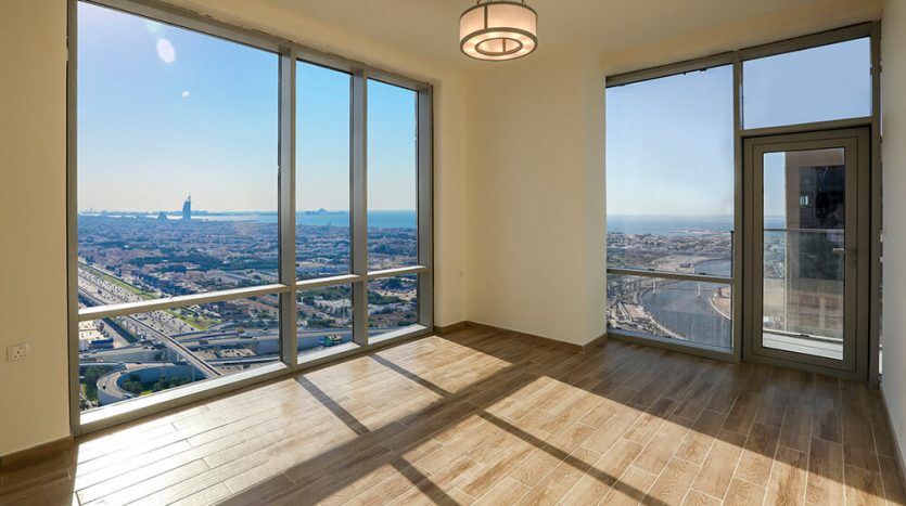 An empty, spacious room with parquet flooring, large windows offering a panoramic view of the rooftops of the Amna Al Habtoor tower under a clear blue sky and a door leading to a balcony.