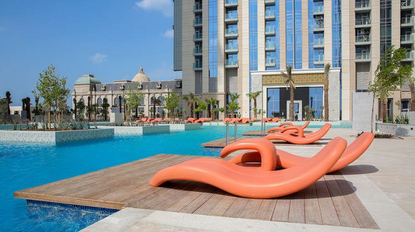Luxury hotel pool area with clear blue water and elegant orange sun loungers on the wooden terrace, surrounded by the Amna Tower and lush palm trees under a brilliant blue sky.