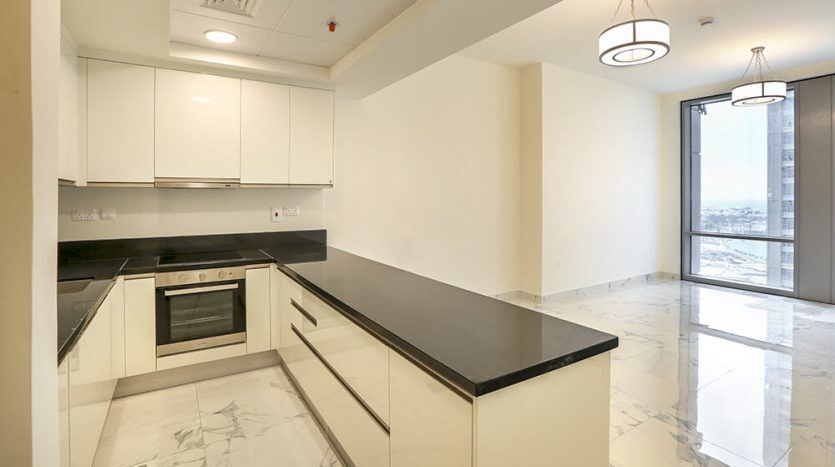 A modern kitchen in the Amna Al Habtoor City tower with white cabinets, black countertops, built-in oven and pendant light, with gleaming marble floors and a window with a view.
