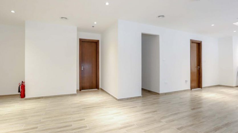 A spacious, modern, empty room in the Amna Tower in Al Habtoor City with light wood flooring, white walls and multiple closed wooden doors. A red fire extinguisher is on the left