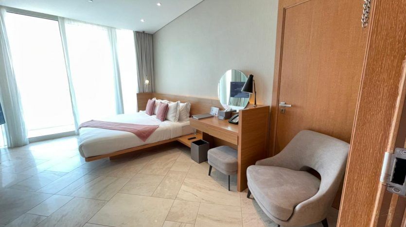 Chambre d'hôtel moderne comprenant un lit double avec des draps blancs, une table de chevet en bois, un fauteuil gris et un grand miroir sur un bureau. La pièce a du carrelage au sol et une grande fenêtre offrant