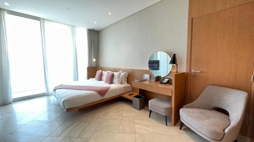 Une chambre d'hôtel moderne Five JVC avec un lit simple, une grande fenêtre, un mobilier en bois comprenant un bureau et une chaise et une décoration neutre.