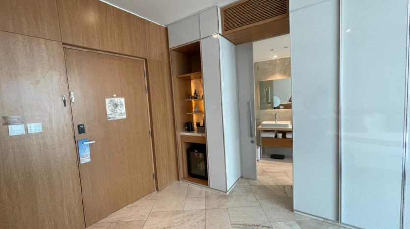 Entrée d'une chambre d'hôtel moderne dotée de portes en bois et d'une armoire à glace à côté d'un minibar visible et d'un aperçu d'une salle de bains avec un grand miroir dans une villa de Dubaï.