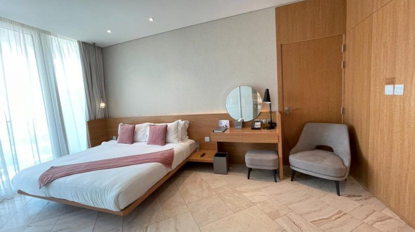 Une chambre d'hôtel moderne avec un grand lit doté d'une literie blanche et rouge, une tête de lit en bois, un bureau en bois adjacent avec miroir, un fauteuil et une baie vitrée avec des rideaux blancs mettant en valeur