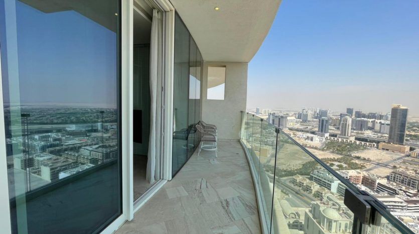 Un balcon de grande hauteur doté d'une balustrade en verre donnant sur un paysage urbain comprenant de nombreux bâtiments dans le quartier Five JVC. Le balcon dispose d'un sol carrelé et d'une seule chaise, et la vue s'étend sous une lumière dégagée.