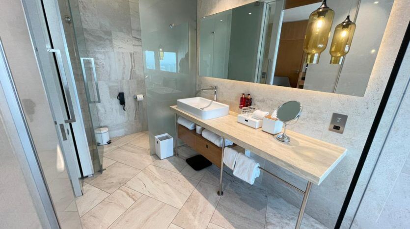Une salle de bains moderne avec un sol en carrelage en marbre, une cabine de douche en verre, une vasque en bois avec un lavabo rectangulaire, un grand miroir et des suspensions Five JVC.