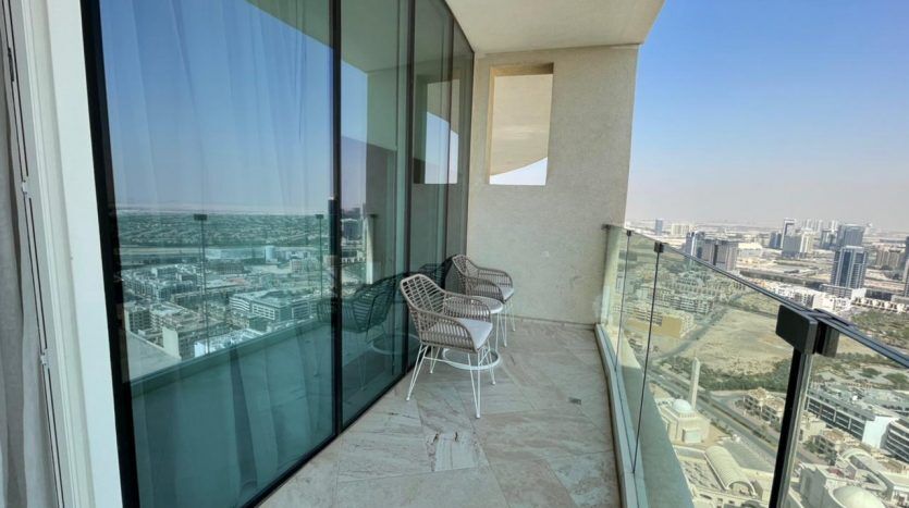 Un balcon en hauteur doté d'une paire de chaises en osier, surplombant un vaste paysage urbain avec des bâtiments modernes sous un ciel clair à Five JVC.