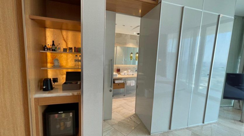 Une chambre d'hôtel moderne dotée d'une étagère intégrée en bois avec minibar et machine à café JVC, ainsi que d'une grande armoire à glace révélant partiellement une salle de bain en marbre.