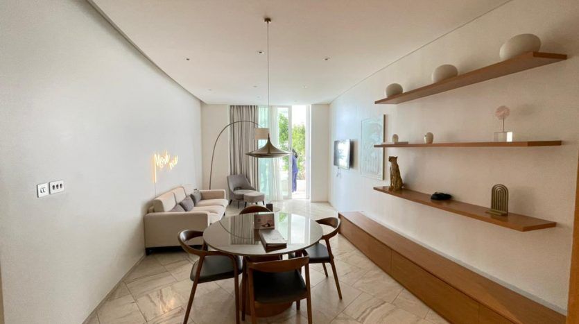 Une salle à manger moderne et minimaliste comprenant une table ronde en verre avec cinq chaises sombres, un canapé sectionnel crème et un décor élégant sous un éclairage doux et naturel.
