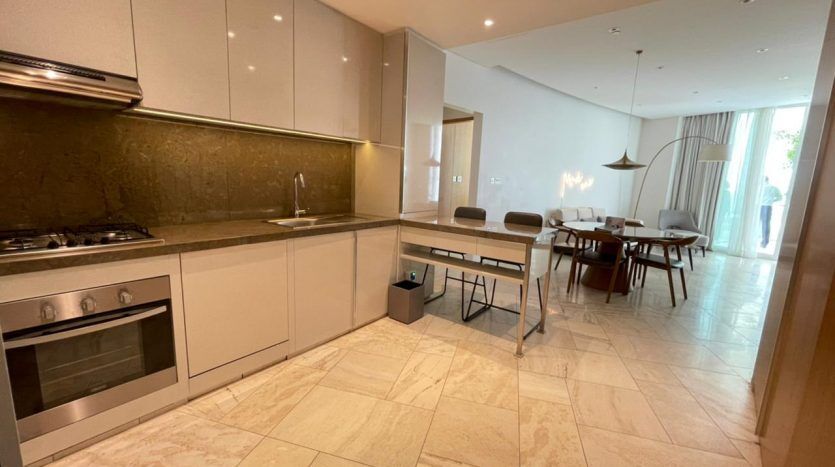Description modifiée : Cuisine et salle à manger modernes avec carrelage beige, armoires beiges élégantes, appareils électroménagers intégrés en acier inoxydable et table à manger Five JVC pour six personnes en arrière-plan. Brillant,
