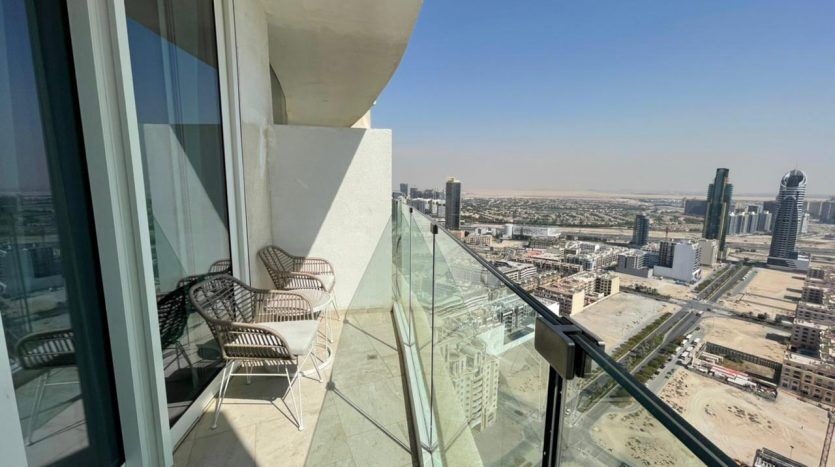 Un balcon en hauteur dans un appartement de Dubaï avec deux chaises donnant sur un paysage urbain avec des gratte-ciel et un vaste terrain ouvert sous un ciel bleu clair.
