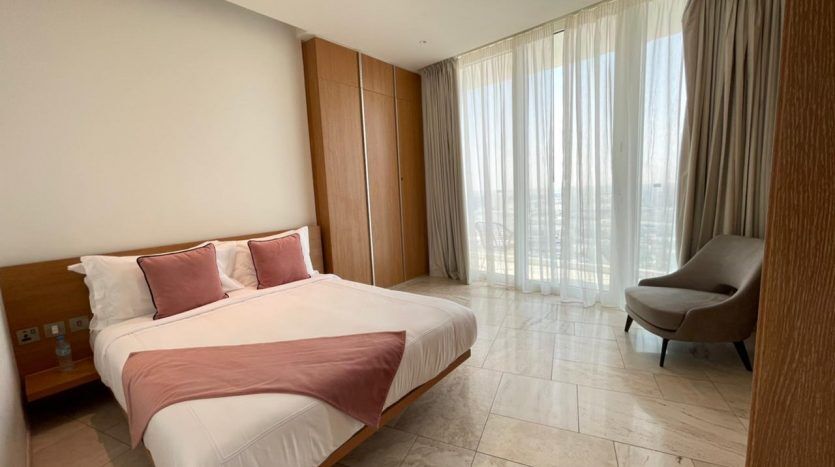 Une chambre moderne avec un grand lit avec une literie blanche et rose, une armoire en bois et un fauteuil beige près d'une baie vitrée avec des rideaux transparents, dotée d'un système audio Five JVC