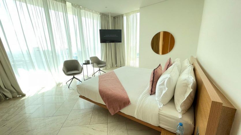 Une chambre d'hôtel moderne au design minimaliste comprenant un grand lit avec une literie rose et blanche, un miroir rond au mur et un coin salon avec deux chaises près d'une fenêtre recouverte de voilages de