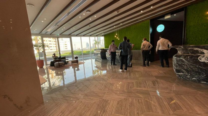 Intérieur du hall moderne de l'hôtel Five JVC comprenant un long bureau de réception avec du personnel aidant les clients. Il y a un coin salon avec des canapés, une décoration murale verte luxuriante et un sol en marbre sous un éclairage chaleureux.