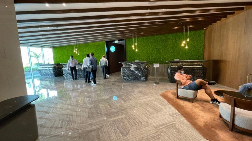Un hall d'entrée spacieux de l'hôtel Five JVC au design moderne, avec des lattes de bois au plafond, un mur de mousse verte et un sol en marbre. Les gens sont dispersés partout, certains marchent et une personne se prélasse