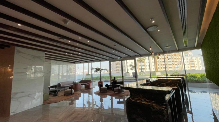 Le hall d'entrée moderne du Five JVC est doté de sols en marbre élégants, de coins salons luxueux et de motifs de plafond à rayures proéminents, donnant sur un paysage urbain à travers de grandes fenêtres.