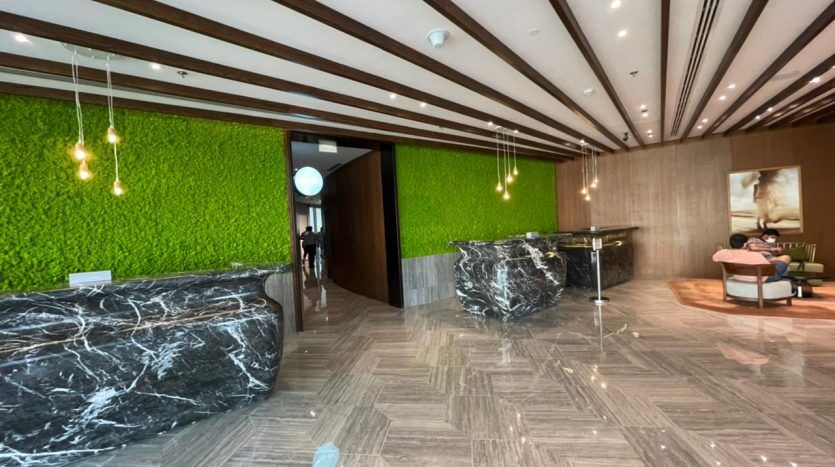 Un espace intérieur d'un bâtiment moderne avec des sols en marbre, des bureaux en marbre noir, des murs de mousse verte vibrante, des lumières suspendues et des poutres en bois au plafond de Five JVC, avec une vue partielle d'une personne dans