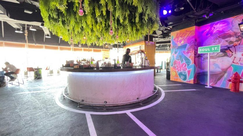 Un bar animé avec un comptoir circulaire, de la verdure suspendue au-dessus et des œuvres d'art murales colorées de style graffiti au Five JVC. Barman et clients sont présents dans un espace intérieur animé et bien éclairé.