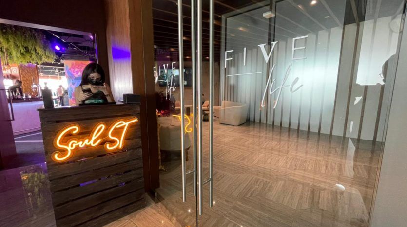 Une zone de réception dotée d'une enseigne au néon indiquant « soul st » sur un bureau en bois avec une personne portant un masque derrière. Portes en verre réfléchissant avec le texte "Five JVC"