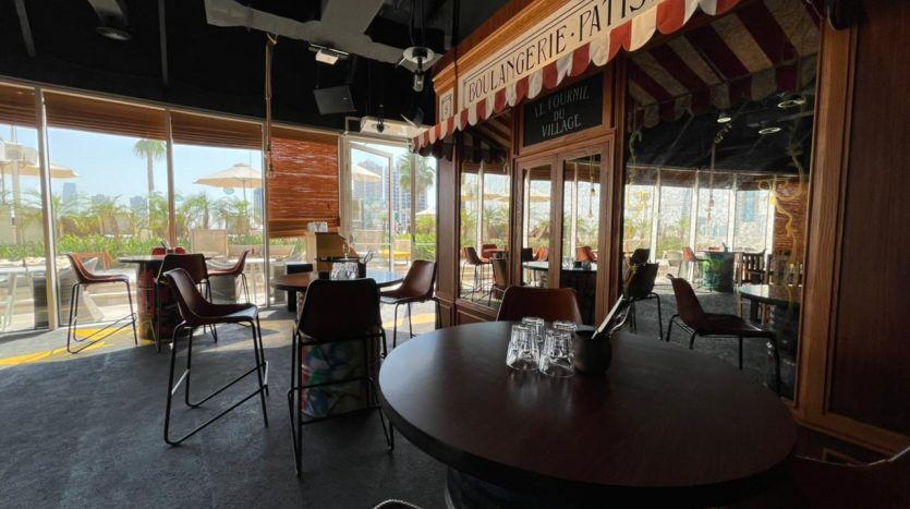 Intérieur d'un café avec un mobilier moderne, de grandes fenêtres laissant entrer la lumière du soleil et une vue extérieure avec des palmiers. Un auvent à rayures rouges indiquant « Five JVC Boulangerie Patisserie