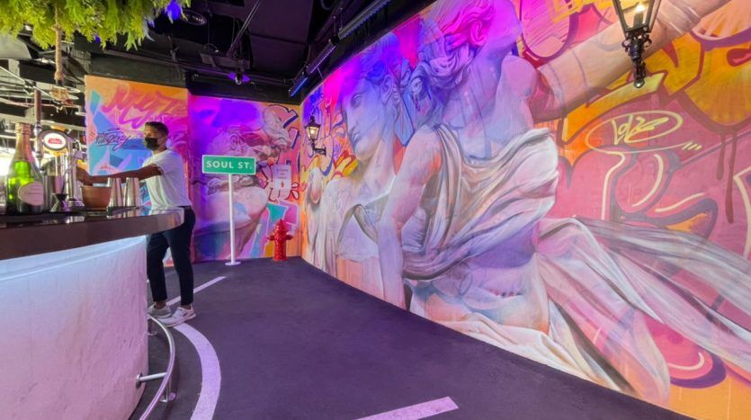 Un bar animé au Five JVC présentant une fresque murale colorée représentant des personnages stylisés sur le mur, un homme debout près du comptoir sous un éclairage d'ambiance et une verdure luxuriante suspendue au-dessus.