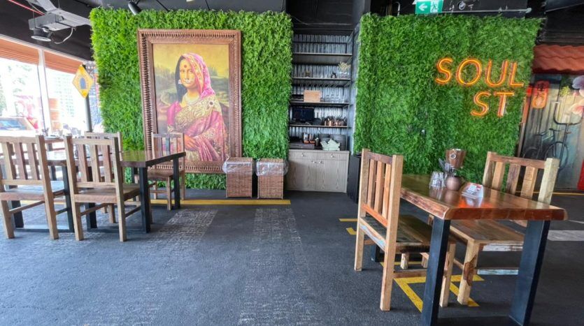 Coin repas intérieur avec tables et chaises en bois, intégré à des murs de verdure artificielle et une grande peinture encadrée représentant une femme en tenue traditionnelle. Un panneau indique "Soul St at Five JVC