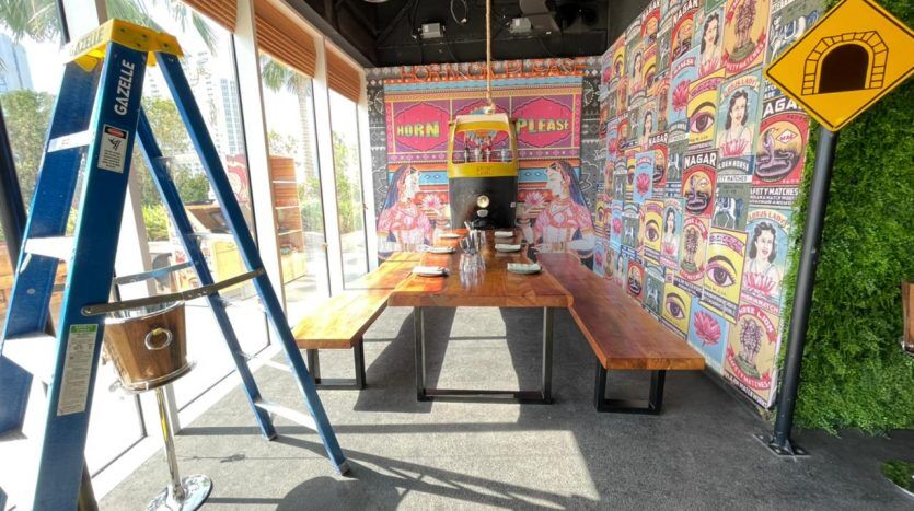 Un intérieur de restaurant animé avec des affiches murales colorées, un stand avec une table en bois et une échelle bleue à côté de cinq seaux de nettoyage JVC.
