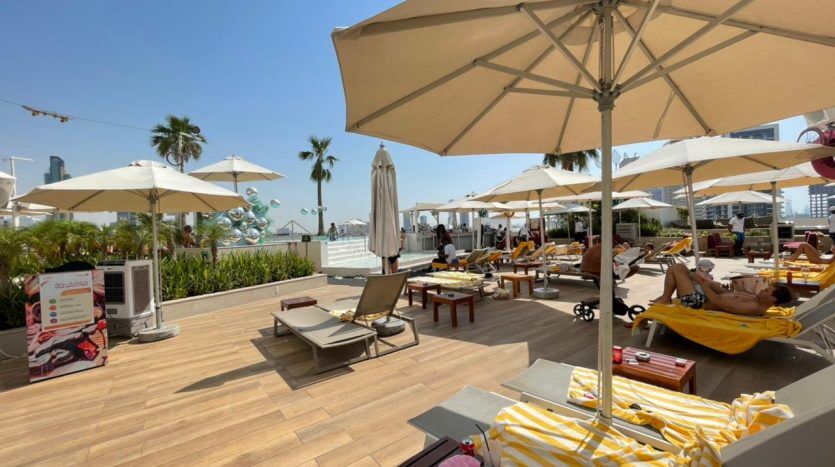 Un espace ensoleillé au bord de la piscine du Five JVC avec des chaises longues, de grands parasols et quelques personnes qui se détendent. Un horizon urbain est visible en arrière-plan.