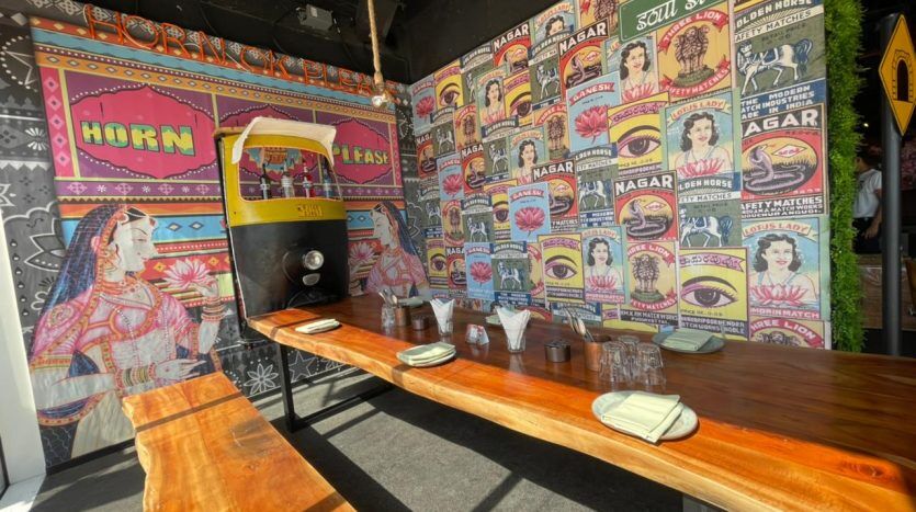 Un intérieur vibrant de restaurant Five JVC sur le thème indien avec un mur mural coloré avec des portraits artistiques, un comptoir de tuk-tuk et des tables communes en bois dressées avec des condiments sous un éclairage naturel.