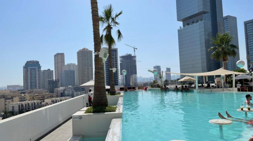 Une piscine à débordement au Five JVC par une journée ensoleillée avec vue sur les toits de la ville, avec des gratte-ciel en construction et un palmier, avec des gens se relaxant dans l'eau.