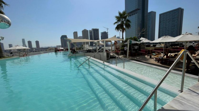 Piscine extérieure de l'hôtel de luxe Five JVC, avec des eaux bleu clair entourées de chaises longues, de parasols et de gratte-ciel sous un ciel ensoleillé.