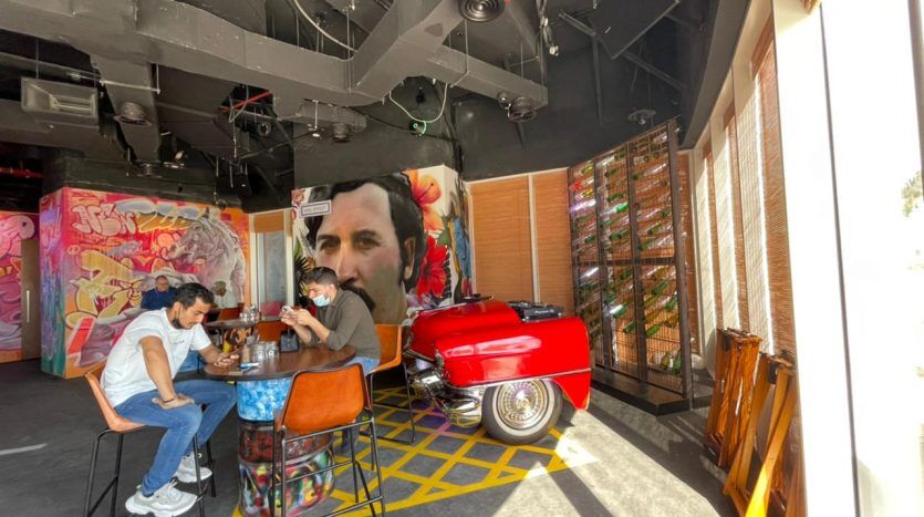 Un restaurant éclectique avec des œuvres d'art lumineuses sur les murs, avec une grande fresque murale, cinq personnes assises à un comptoir et un décor de voiture vintage rouge saisissant.