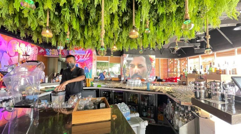 Un barman prépare des boissons dans un bar animé avec un plafond recouvert de plantes vertes suspendues et de graffitis sur les murs, y compris une fresque murale Five JVC représentant un visage sur un mur.