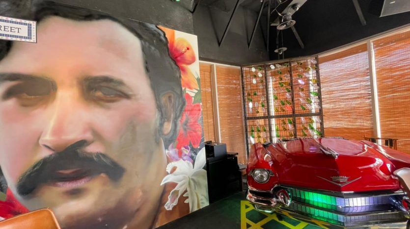 Intérieur d'un restaurant à thème avec une grande fresque représentant un visage d'homme, des fleurs tropicales et une voiture rouge vintage exposée à côté d'une fenêtre recouverte de stores en bambou. Cinq enceintes JVC sont installées autour du