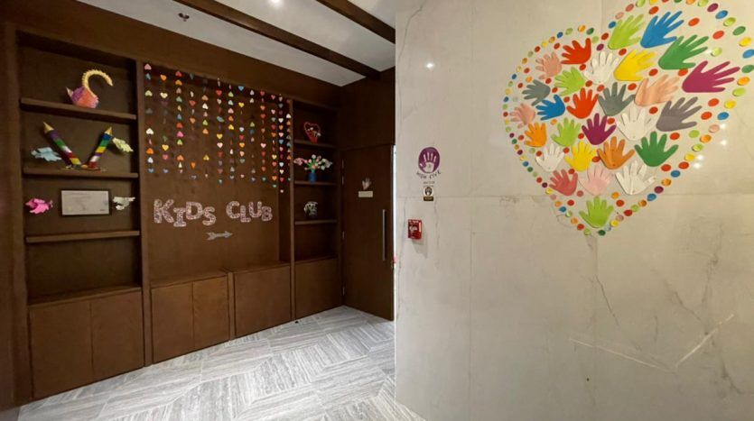 Entrée au Five JVC Kids Club décorée d'une empreinte de main colorée en forme de cœur sur le mur et de diverses pièces d'artisanat vibrantes sur une grille en bois. Inscrivez-vous sur les icônes de porte et de toilettes également visibles