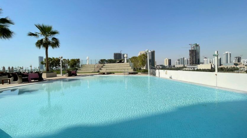 Une piscine extérieure sur le toit du Five JVC avec une eau bleu clair, entourée de chaises longues et de palmiers, surplombant les toits de la ville sous un ciel clair.