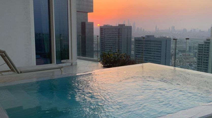 Une piscine à débordement sur le toit du Five JVC surplombe les toits de la ville au coucher du soleil, avec des nuances d'orange et de rose dans le ciel, se reflétant sur la façade vitrée du bâtiment et l'eau de la piscine.