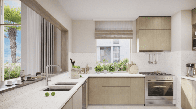 Cuisine moderne avec armoires en bois clair, comptoirs blancs et appareils électroménagers intégrés en acier inoxydable à Tical Al Furjan. Une verdure abondante et la lumière naturelle provenant de grandes fenêtres rehaussent l'espace.