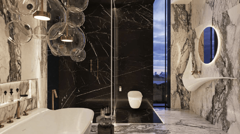 Salle de bains luxueuse dans un appartement de Dubaï avec sols et murs en marbre, comprenant une baignoire autoportante, des luminaires modernes, une cabine de douche en verre et un miroir ovale unique. Un éclairage élégant met en valeur l'élégance