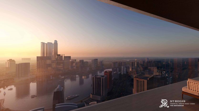 Sunset view of a Dubai cityscape featuring a river, modern skyscrapers and several buildings, seen from a high balcony. Bright orange tones are reflected on the water and buildings.