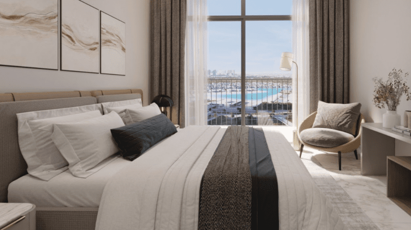 A modern bedroom in a Dubai villa with a large bed, beige and black blankets and plenty of pillows. A desk and chair are to the right, next to a window with a view of the city.