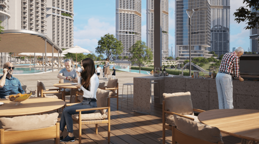 Outdoor rooftop dining area with people socializing in Dubai. A man grills food while others sit at tables conversing, surrounded by modern buildings and a swimming pool.
