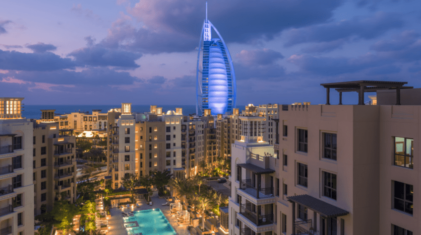 View of the Burj Al Arab from the balcony of an apartment at dusk, surrounded by other buildings and illuminated pools, under a slowly fading sky.