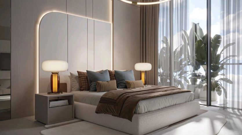 A modern bedroom with a large Legado Prescott bed, flanked by two bedside tables, each supporting a lamp with an amber-colored cylindrical base and white shade. The room features an elegant, light-colored headboard, picture windows with sheer curtains and potted plants.