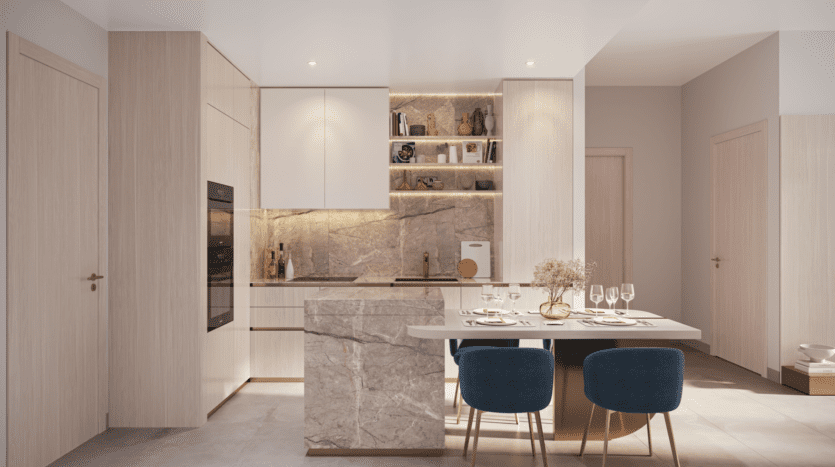 A modern kitchen with light wood cabinets, built-in oven and marble countertops embodies the Legado Prescott style. A small dining area includes a marble table with plates and glasses, surrounded by blue chairs. Subtle lighting enhances the clean, contemporary design.