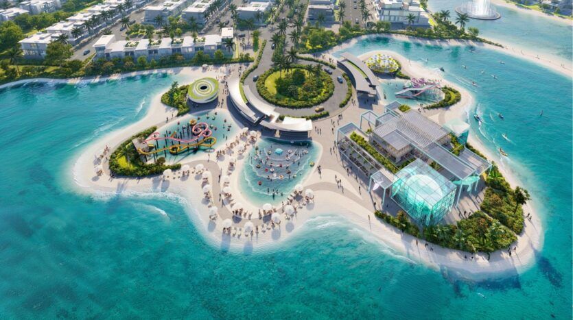 Vue aérienne de DAMAC Islands 2, une île de villégiature dynamique avec des piscines, des toboggans aquatiques, des espaces de détente, des parasols, des bâtiments modernes et des palmiers entourés d'une eau claire et turquoise et de plages de sable.