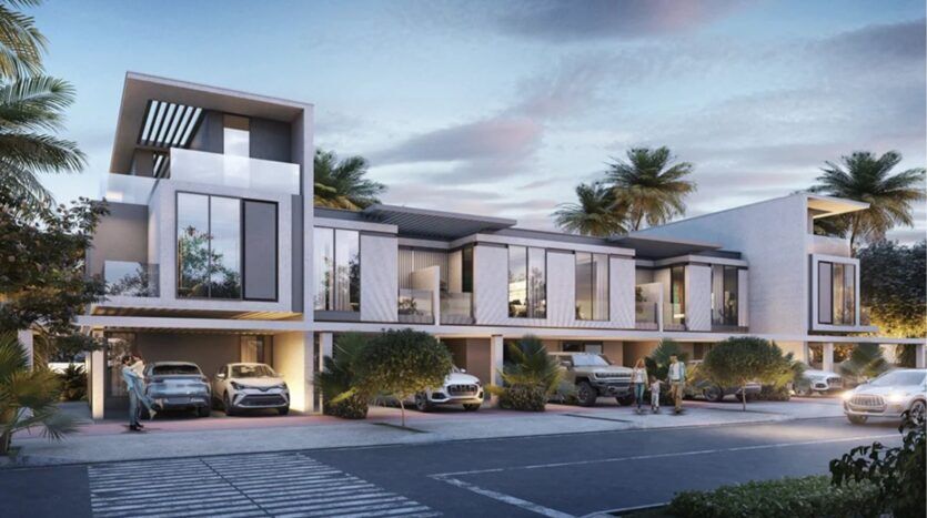 Les maisons de ville modernes de deux étages sur Damac Islands 2 présentent de grandes baies vitrées, des carports avec plusieurs voitures garées, des palmiers et des personnes debout à l'extérieur, le tout sous un ciel partiellement nuageux le soir.