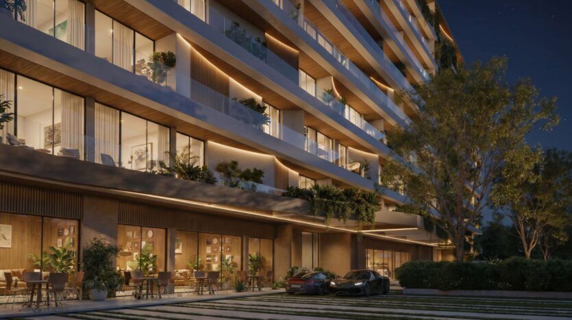 Lunaya Terraces : un immeuble d'appartements moderne vu de nuit, avec un éclairage chaleureux, de grandes fenêtres, des balcons agrémentés de plantes, des coins salon en plein air au rez-de-chaussée et deux voitures garées devant, le tout entouré de verdure et d'arbres.